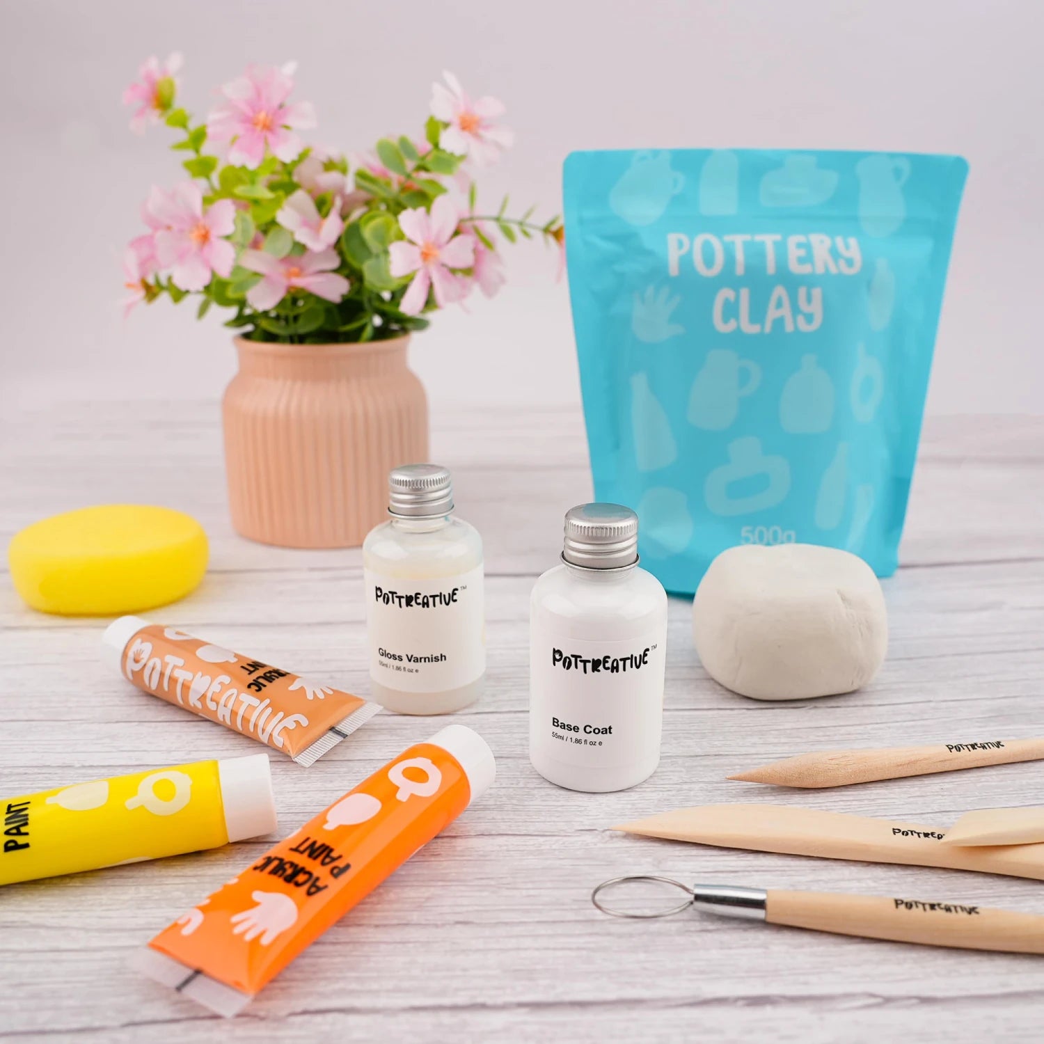 A close-up of the Pottreative air dry clay kit materials, featuring clay, base coat, gloss varnish, acrylic paints, and wooden sculpting tools.